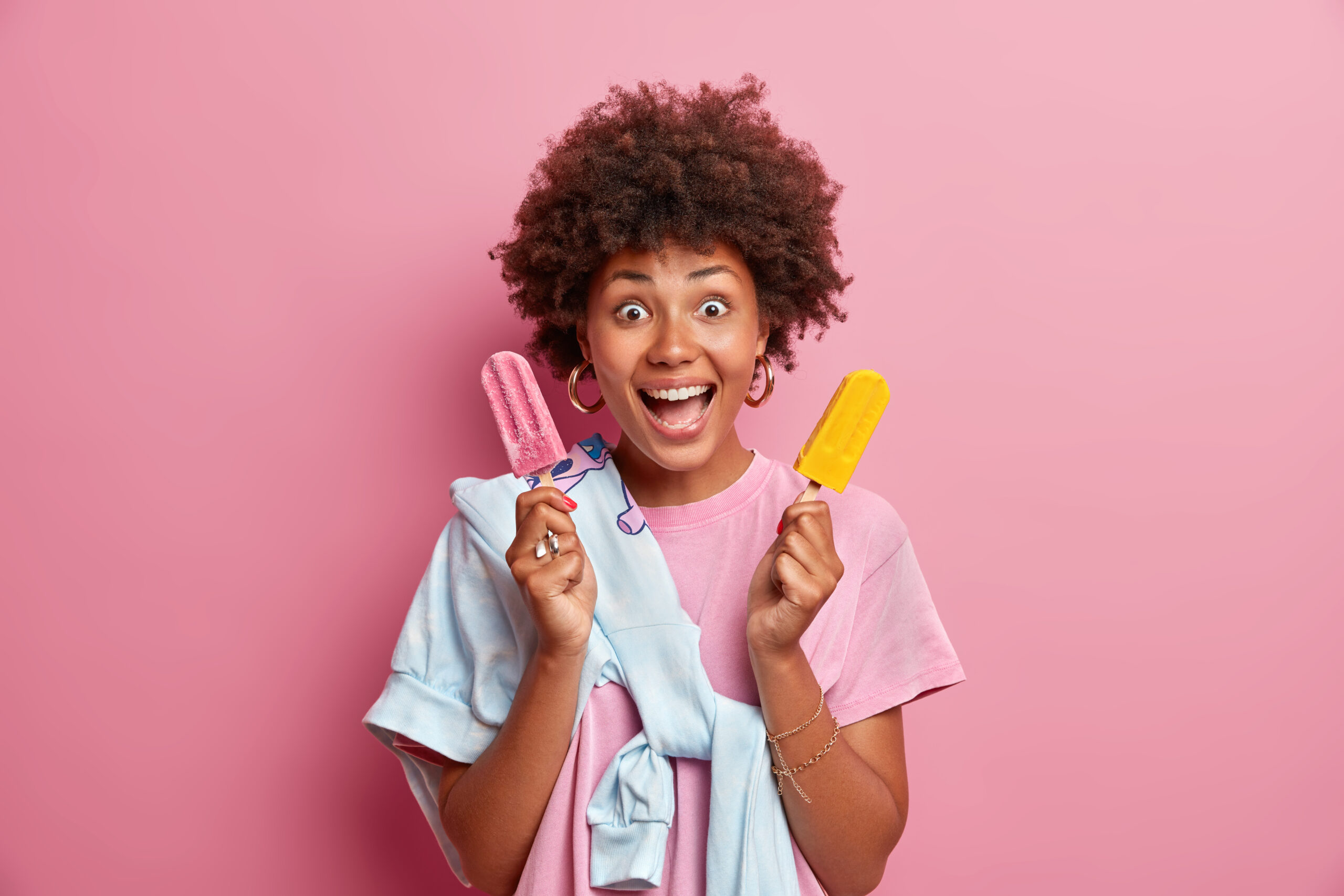 Glad dark skinned woman holds two cold ice creams, keeps mouth opened, treats herself with delicious dessert, enjoys pleasant taste, poses against rosy background. Summer vacation, happiness